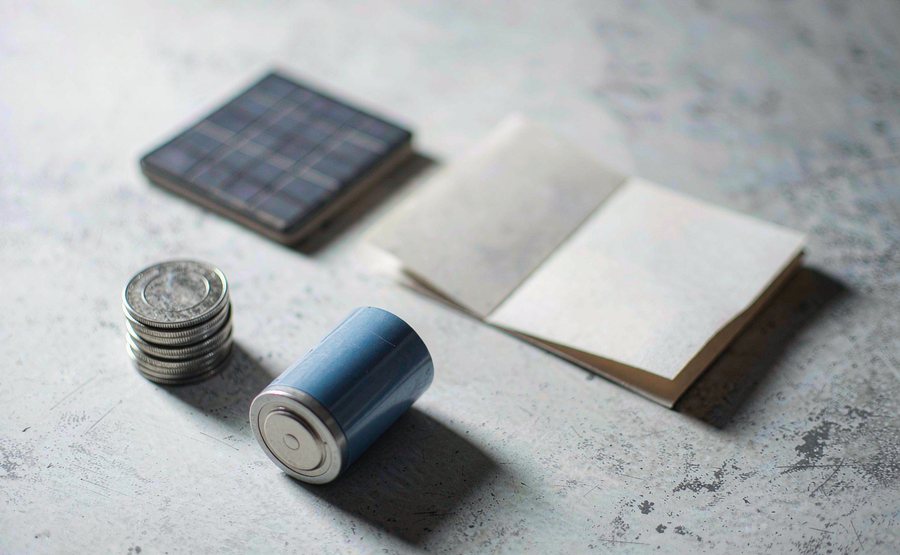 A stack of coins, a rechargeable battery cell, a small solar panel and a folded bill on a grey desk