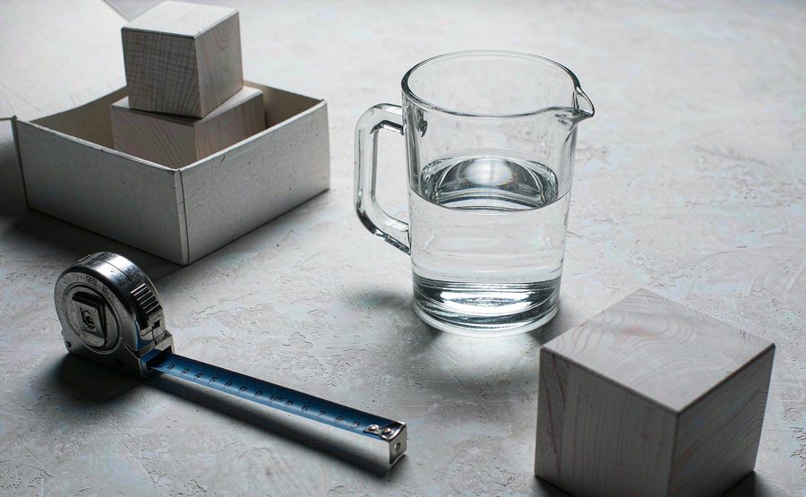 A tape measure, a glass jug of water, wooden blocks and an open cardboard box on a grey desk