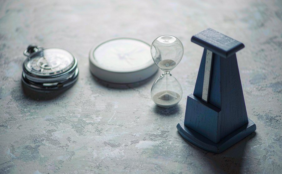 A chrome stopwatch, a glass hourglass, a blank clock face and a metronome on a grey desk