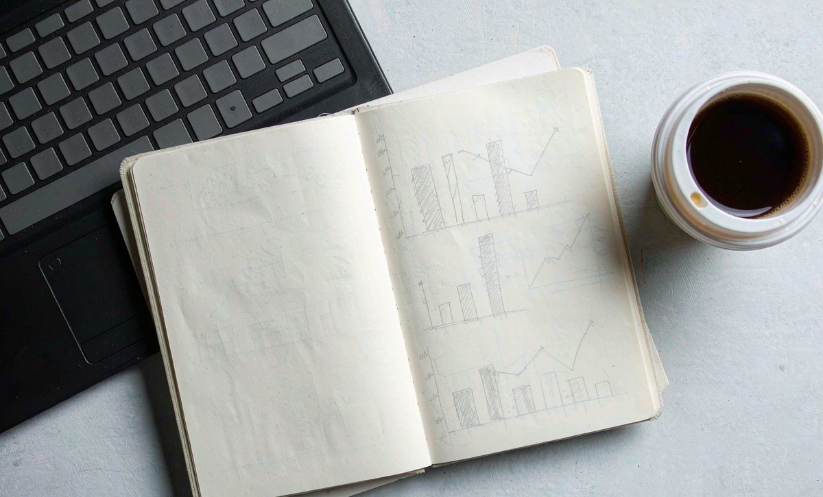 An open notebook with hand-drawn bar and line charts beside a laptop and a cup of coffee