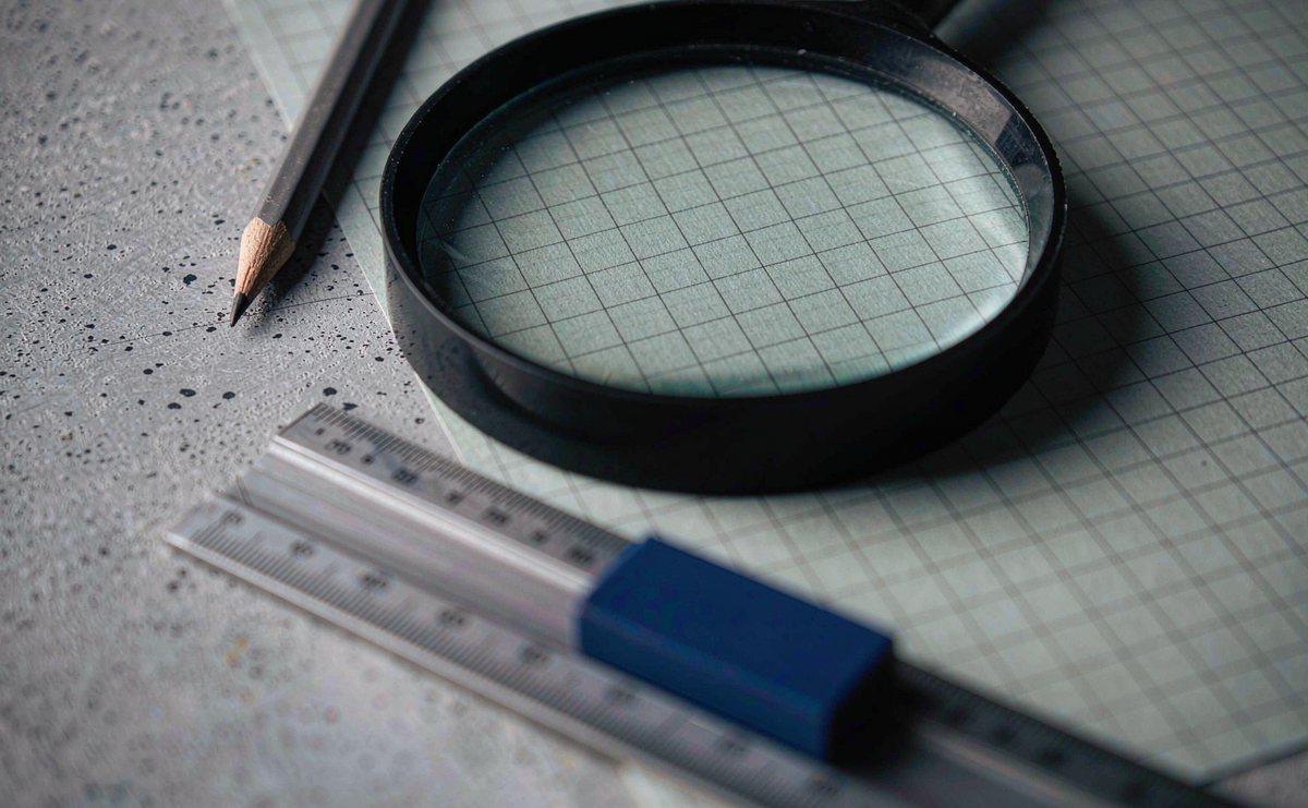 A magnifying glass resting on graph paper beside a sharpened pencil and a short steel ruler